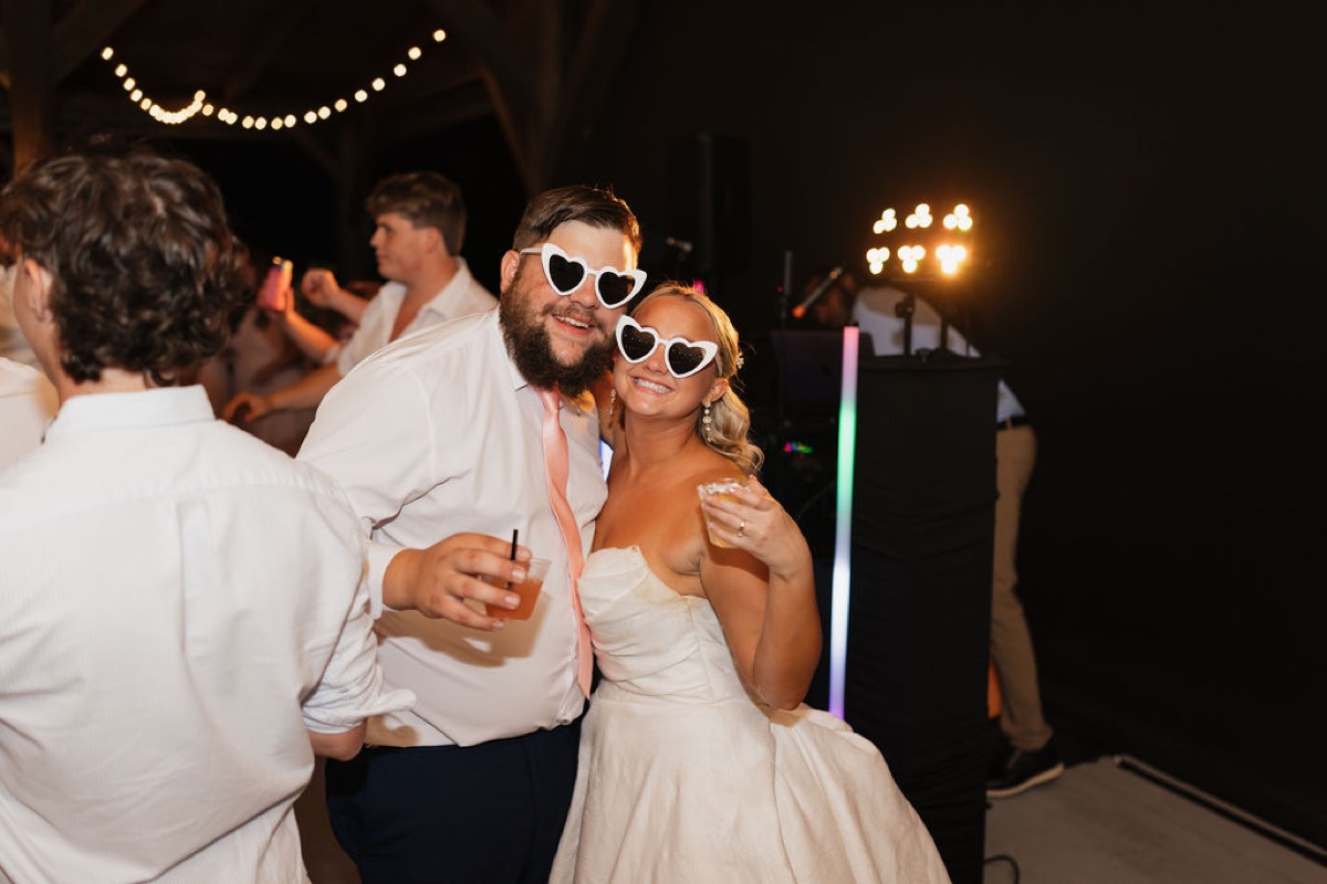 Lilly & Jackson in heart sunglasses toast with drinks under string lights at their Covey Creek reception