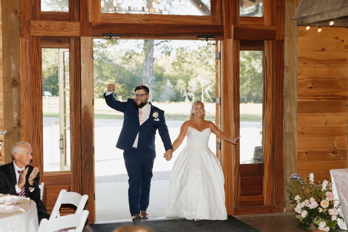 Lilly & Jackson enter Covey Creek reception through barn glass doors as Jackson raises a triumphant fist