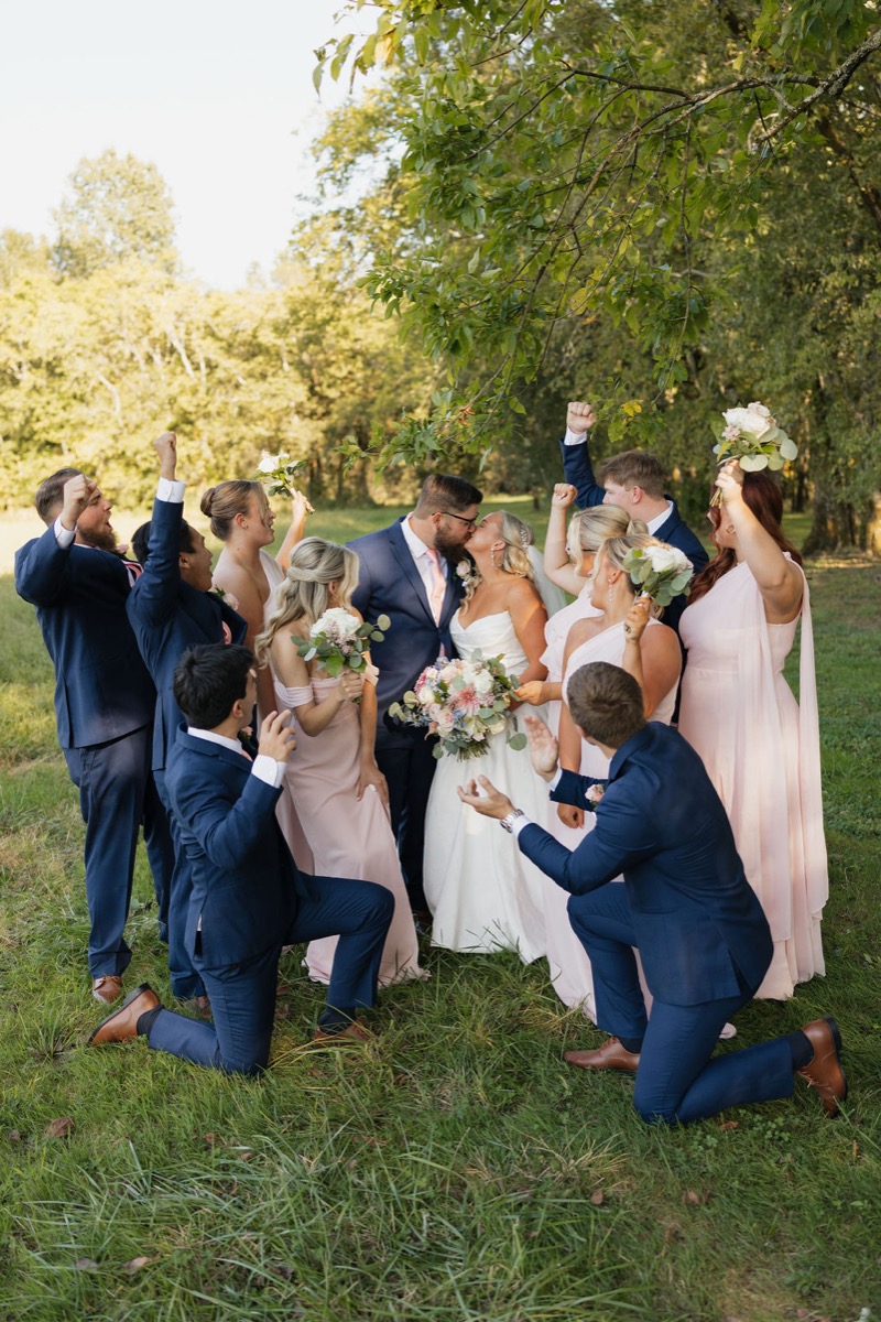 Lilly & Jackson kiss as navy-suited groomsmen and blush bridesmaids cheer in a sunny field at Covey Creek