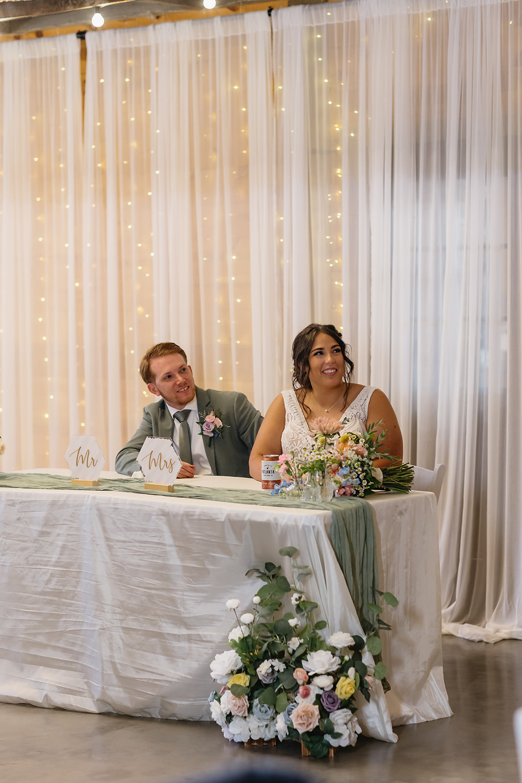 Meredith & Benjamin smile at their sweetheart table with Mr & Mrs signs in the barn at Covey Creek
