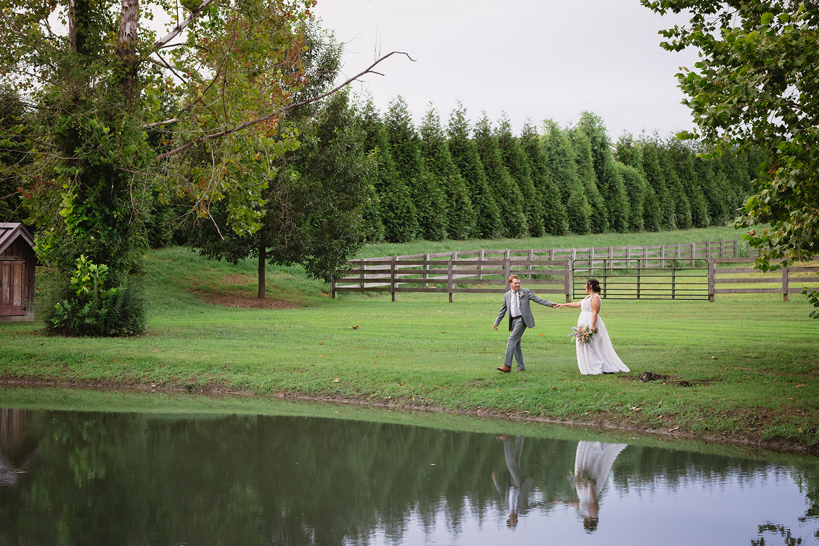 Meredith & Benjamin walk hand in hand beside the Pond with rail fence and trees at Covey Creek