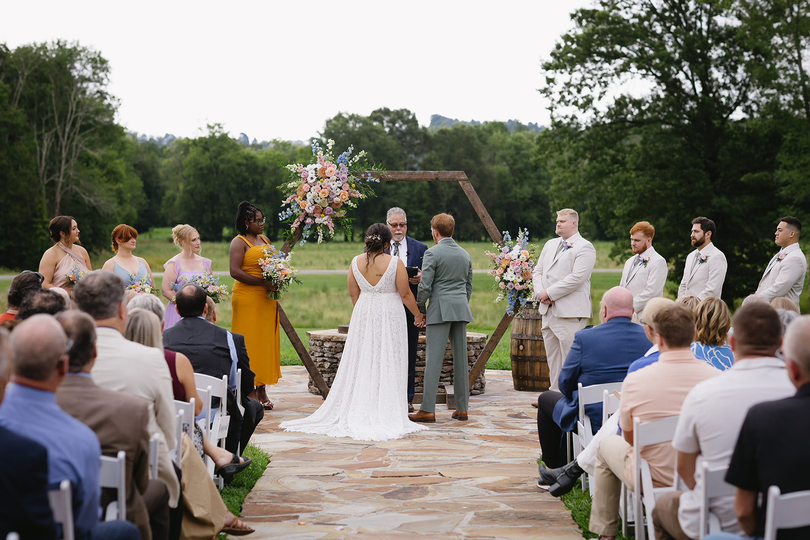 Meredith & Benjamin face the officiant under the Patio hexagon arch as guests watch at Covey Creek