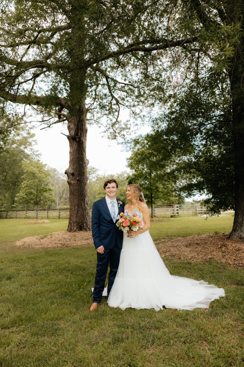 Morgan & Caden pose on the green lawn at Covey Creek, her colorful bouquet bright against fence and trees