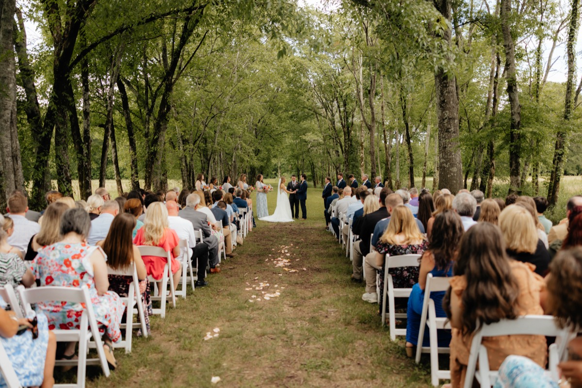 Guests fill white chairs along Morgan & Caden’s petal-strewn Tunnel of Love aisle at Covey Creek