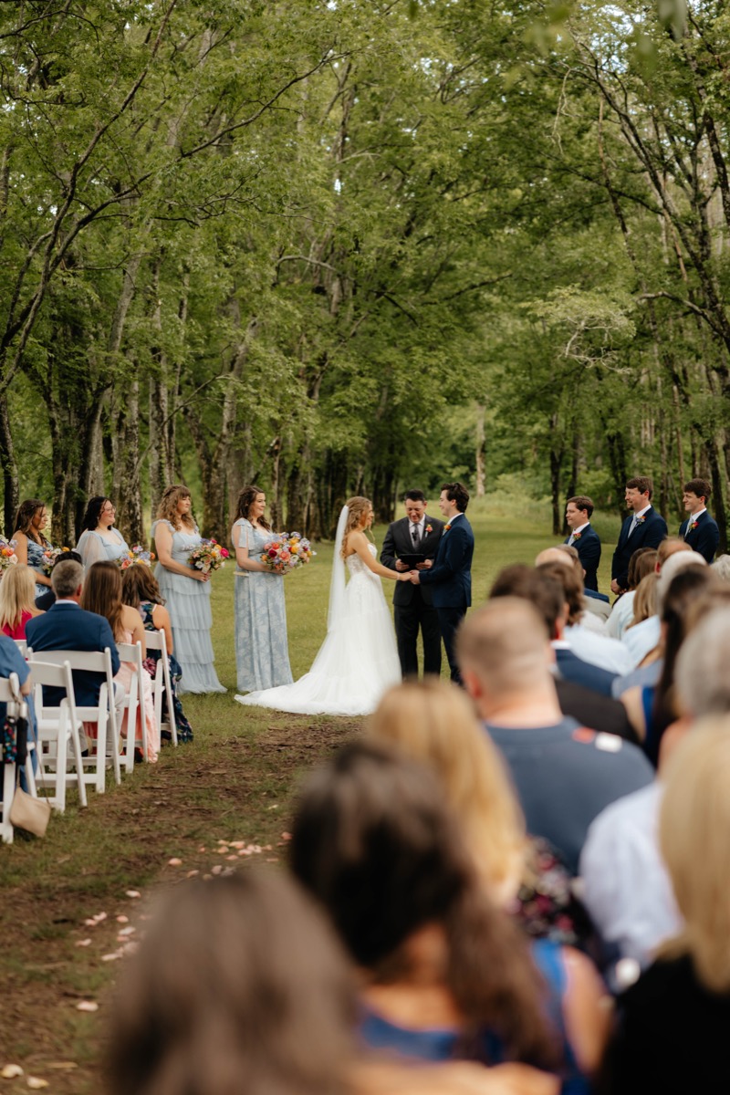 Morgan & Caden hold hands along Covey Creek’s Tunnel of Love aisle as guests and blue bridesmaids look on
