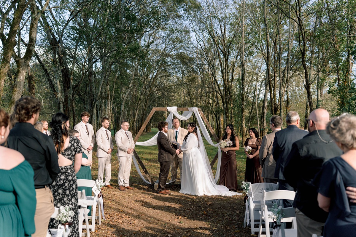 Guests watch Peyton & Jackson exchange vows under a hexagonal wooden arbor in the woods at Covey Creek.