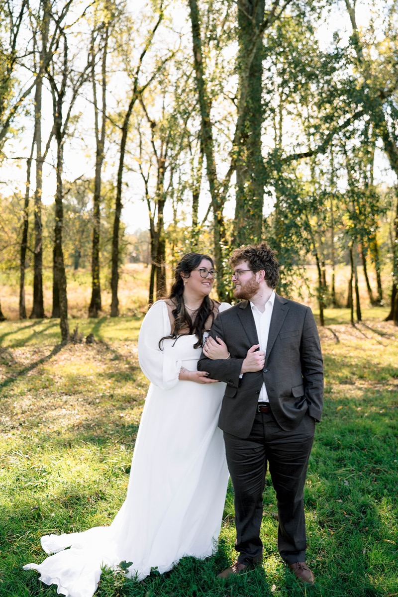 Peyton & Jackson stand arm-in-arm in a sunlit grassy clearing among tall trees at Covey Creek.