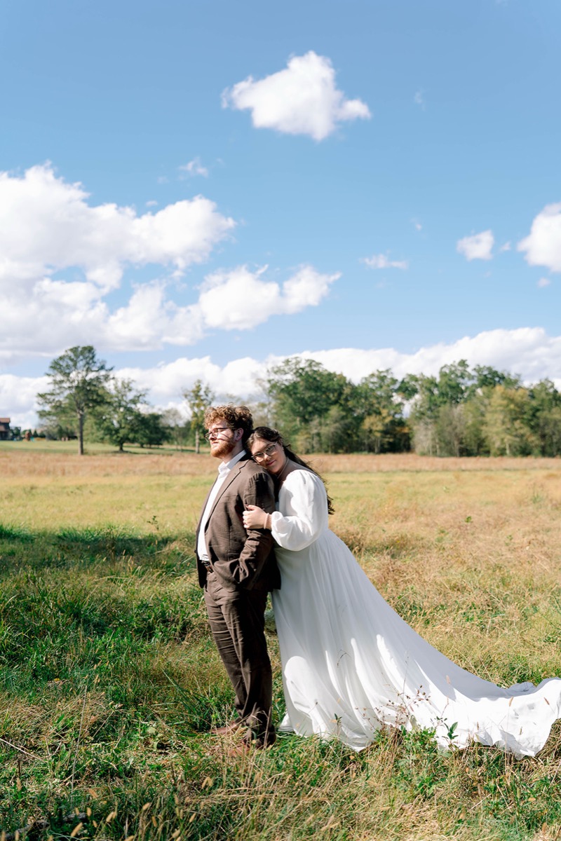 Peyton & Jackson stand in tall grass as she leans on his shoulder under blue sky at Covey Creek.