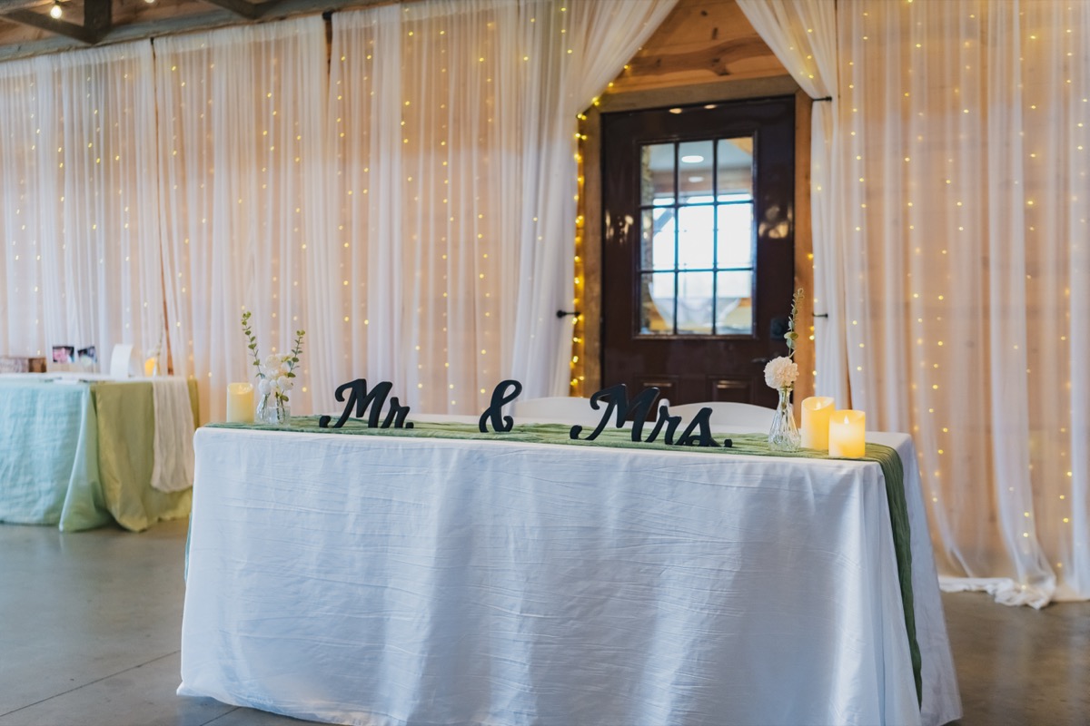 Rae & David sweetheart table with Mr & Mrs sign, candles, and fairy-lit curtains at Covey Creek indoor reception