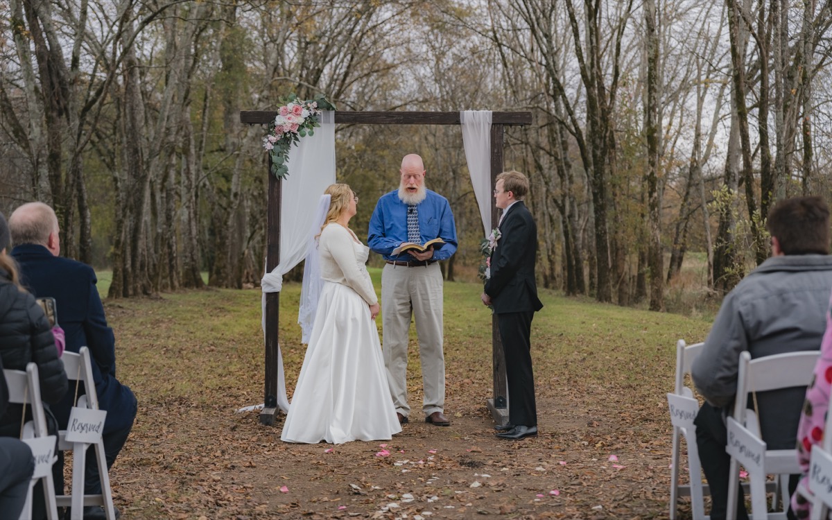 Rae & David stand with their officiant under a white-draped arbor at Covey Creek Tunnel of Love ceremony