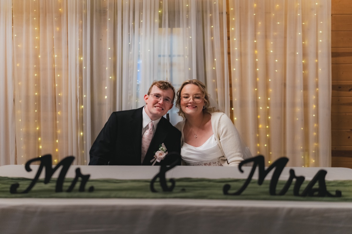 Rae & David smile at their sweetheart table behind a Mr & Mrs sign at Covey Creek indoor reception