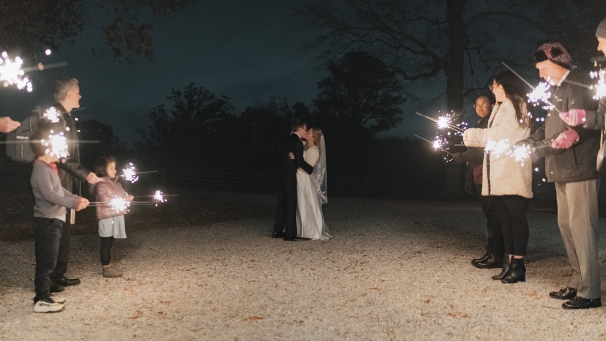Rae & David kiss as guests hold sparklers along a gravel path at Covey Creek nighttime send-off