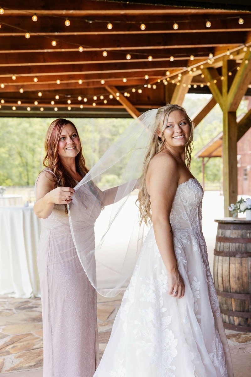 Raven smiles as her mother holds her veil under pavilion string lights at Covey Creek for Raven & Jackson
