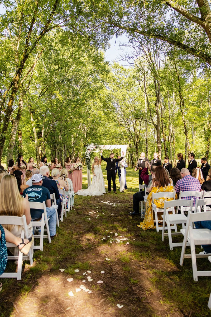 Raven & Jackson cheer with arms raised under their floral arbor as guests watch at Covey Creek