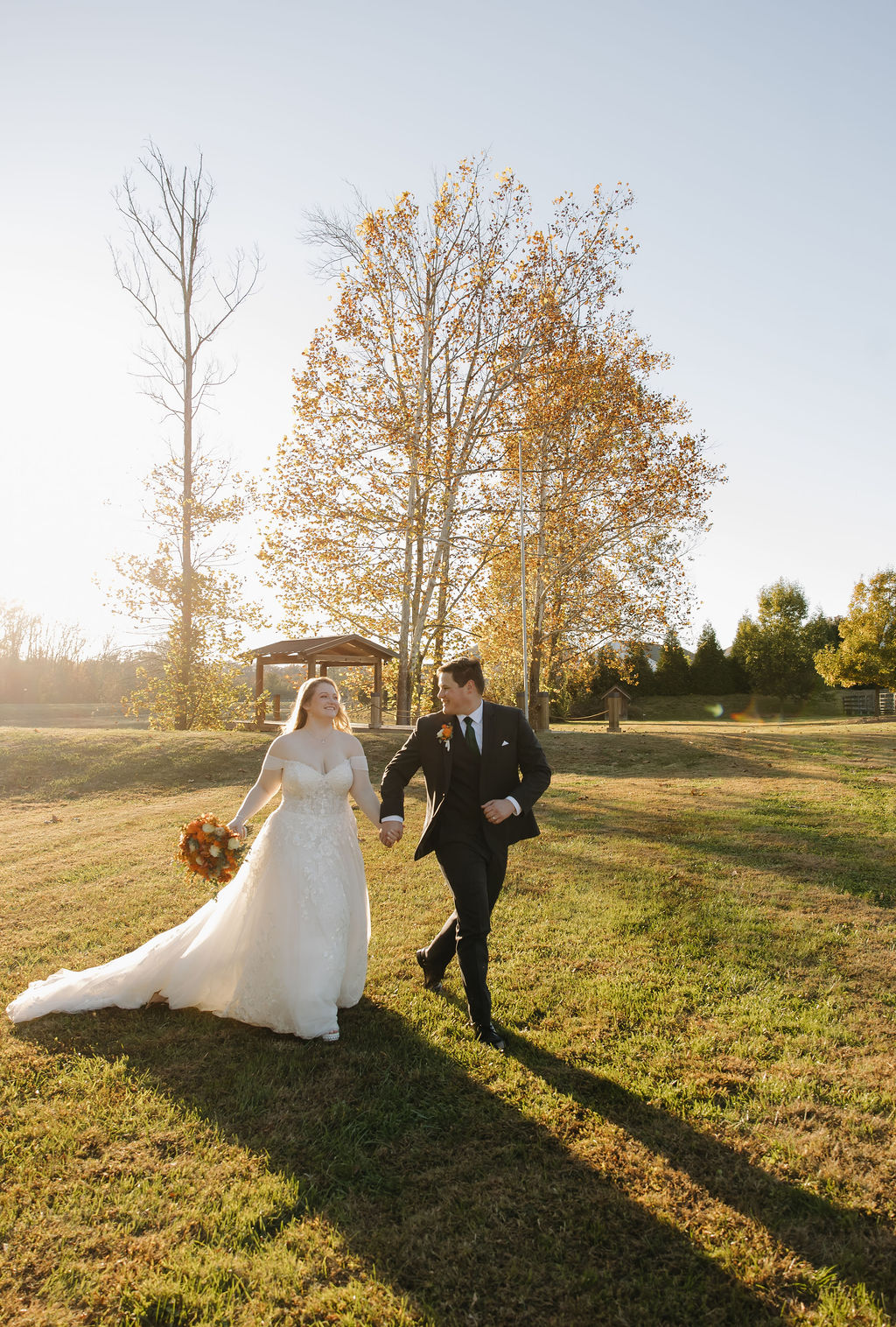 Rebecca & Troy walk hand in hand across a sunlit grassy field beneath golden fall trees at Covey Creek.