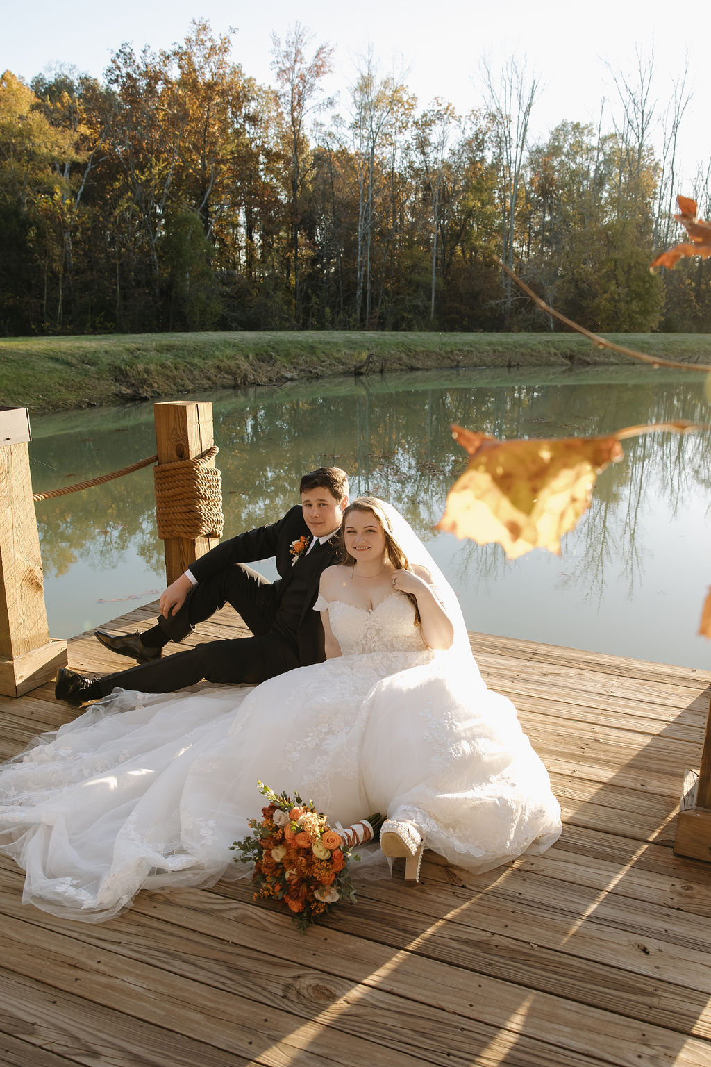 Rebecca & Troy sit on a weathered dock by the pond, autumn trees behind them, smiling at Covey Creek.
