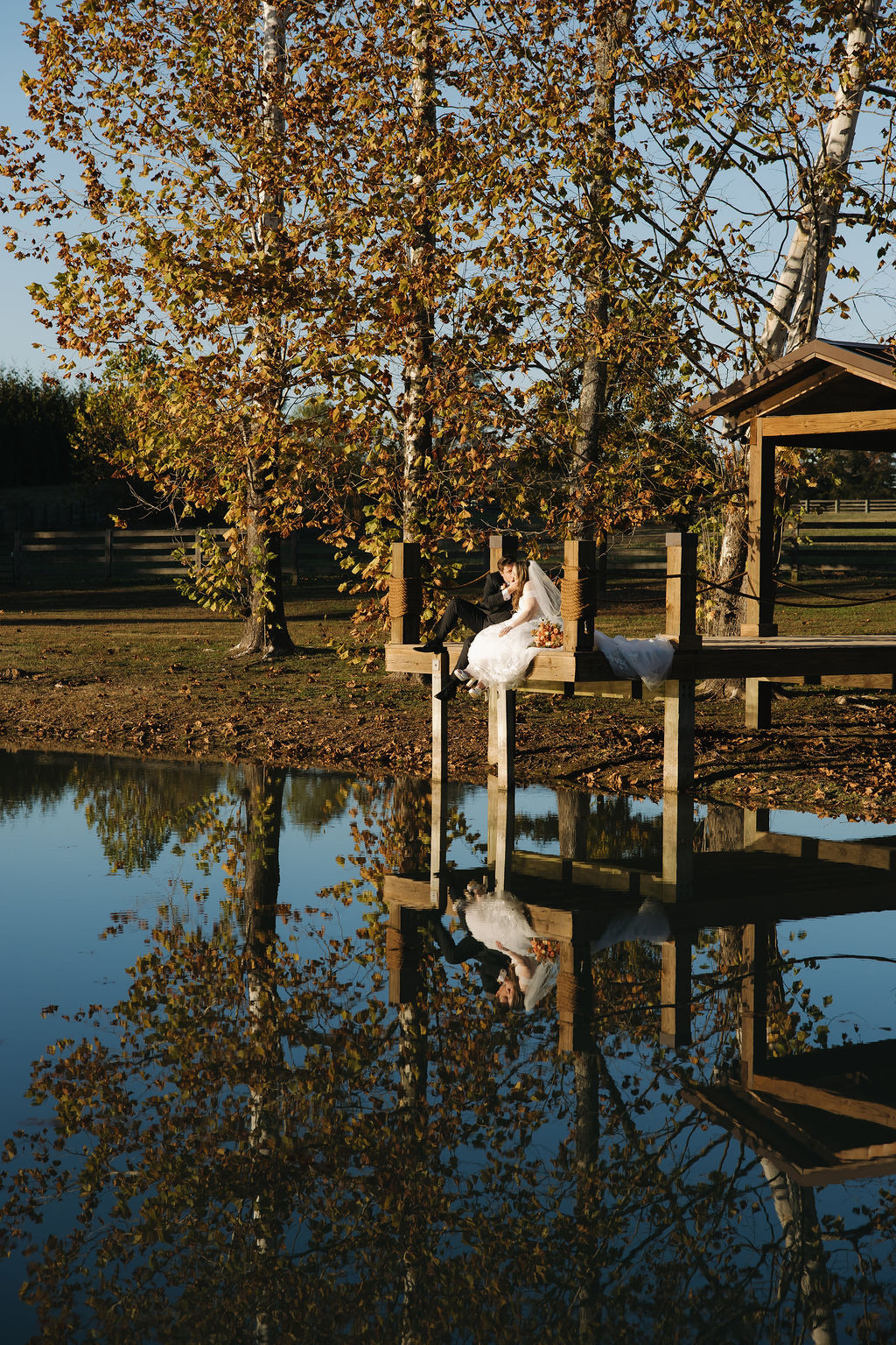 Rebecca & Troy sit on Covey Creek’s pond dock, their reflection mirrored in calm water under golden autumn trees.