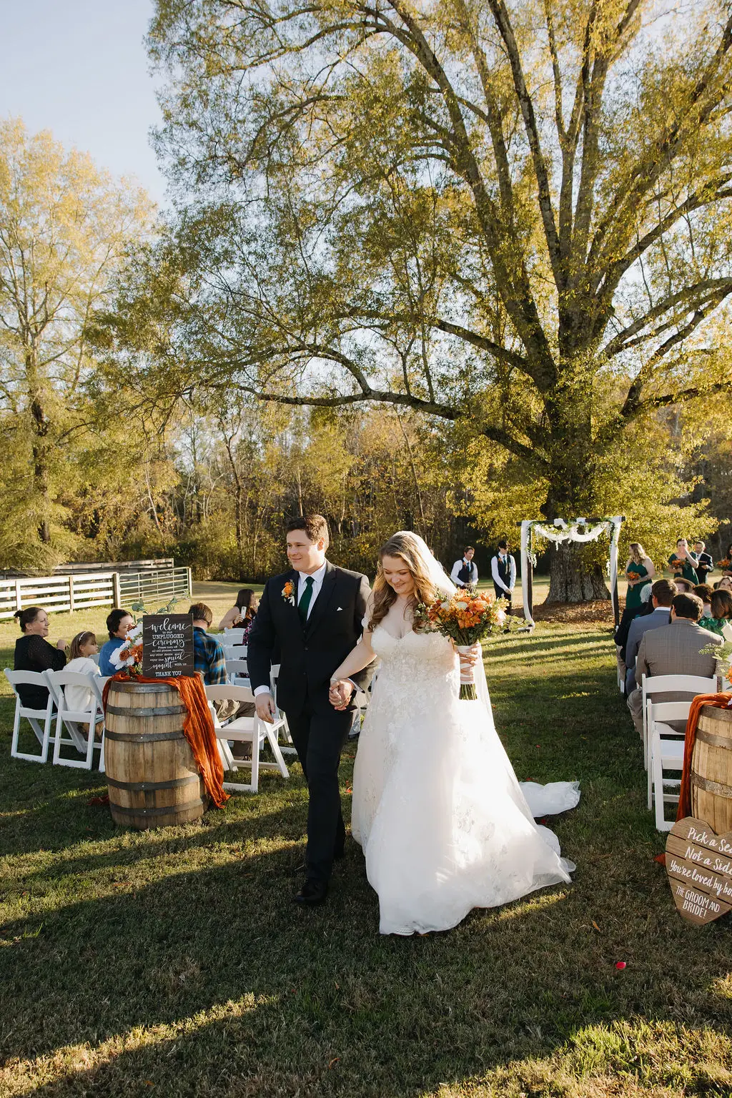 Rebecca & Troy walk the grassy aisle after their outdoor autumn ceremony beneath a large tree at Covey Creek.