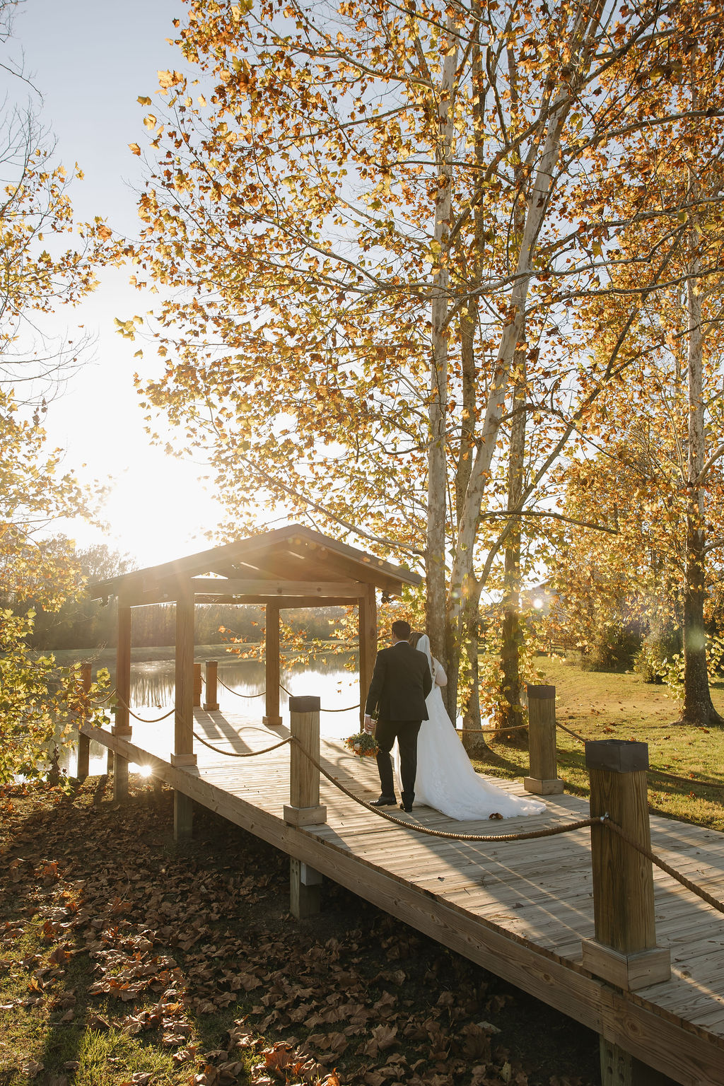 Rebecca & Troy walk the wooden pond dock toward a small pavilion amid golden fall trees at Covey Creek.