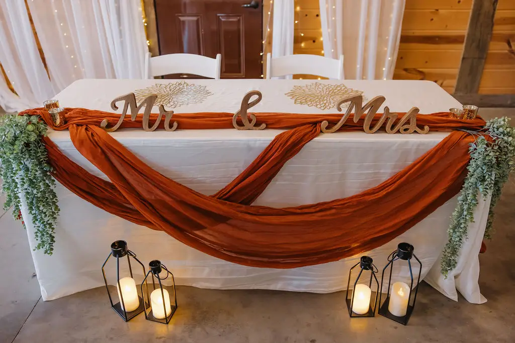 Rebecca & Troy’s sweetheart table with Mr. & Mrs. letters, rust draping, and candle lanterns indoors at Covey Creek.