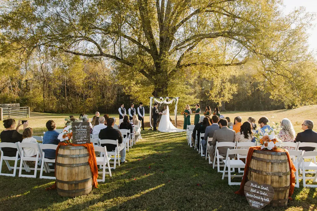 Rebecca & Troy stand at a tree-shaded arbor as guests fill white chairs along the grassy aisle at Covey Creek.