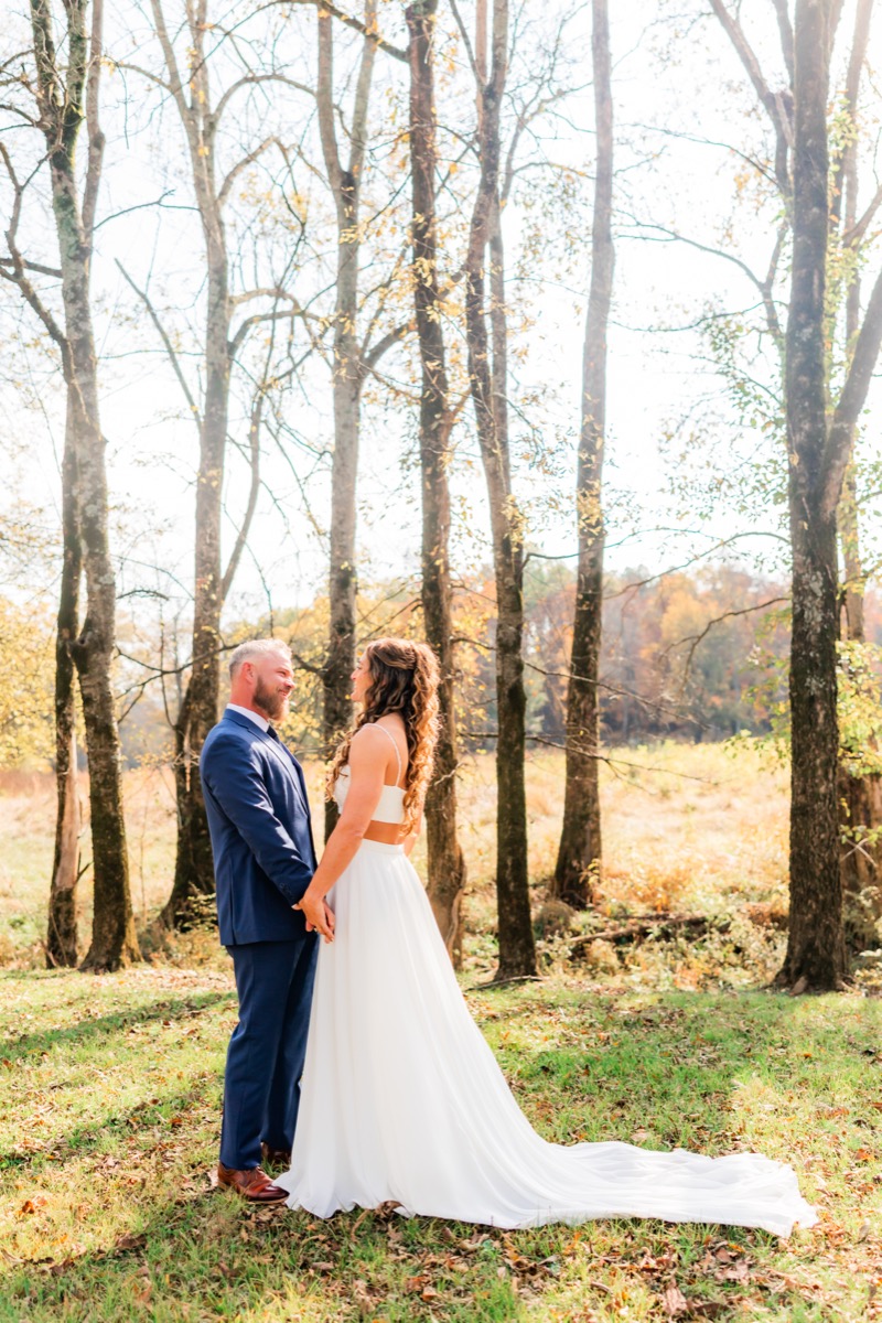 Sarah & Adam hold hands face-to-face among tall trees in a sunlit autumn clearing at Covey Creek