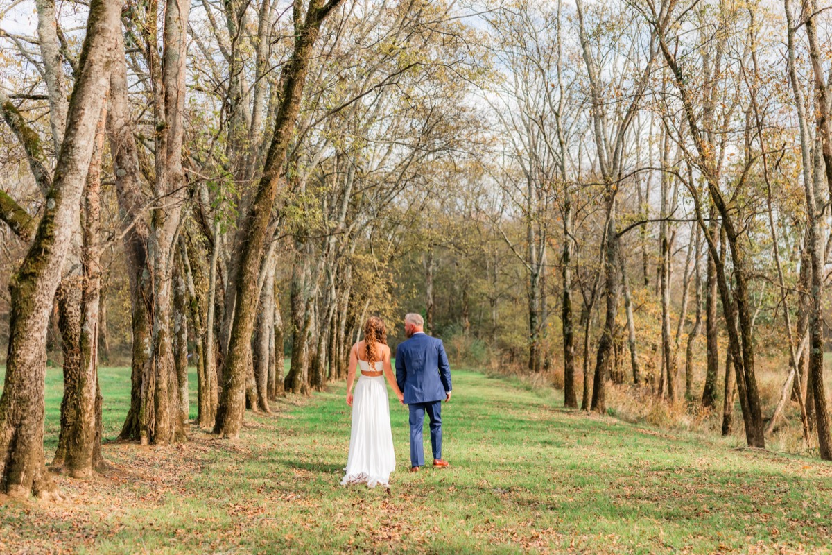 Sarah & Adam walk hand-in-hand down a leaf-strewn tree-lined path on their Covey Creek wedding day