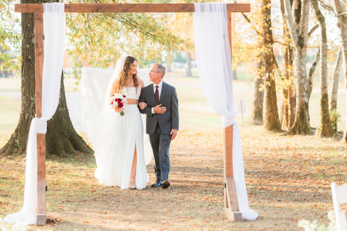 Sarah & Adam's bride walks the aisle under a white-draped wooden arbor with her father at Covey Creek