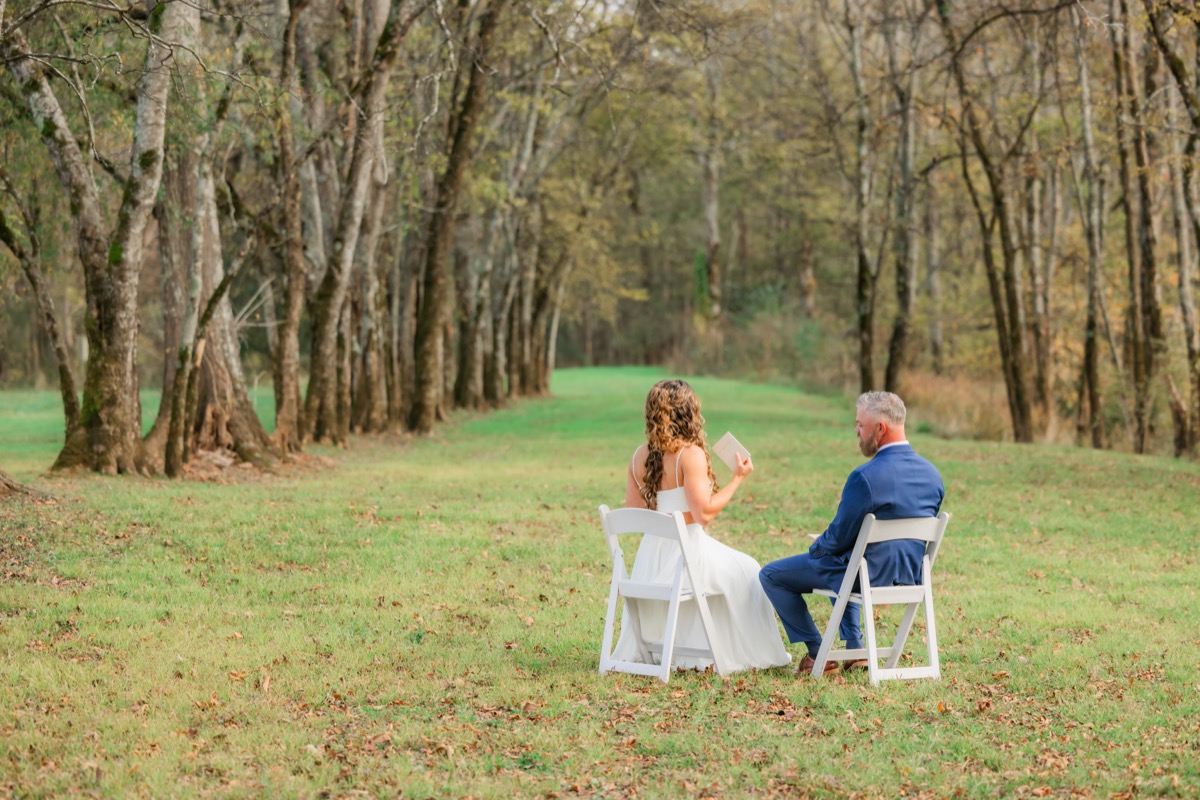 Sarah & Adam sit in white chairs exchanging words along a tree-lined autumn field at Covey Creek
