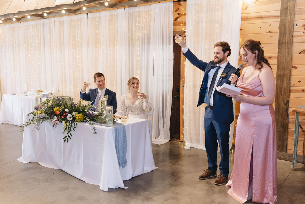 Shay & Jacob raise glasses at their sweetheart table while a bridesmaid toasts under string lights at Covey Creek.