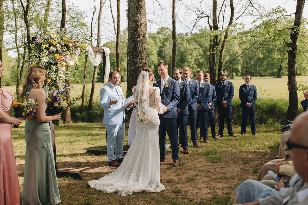 Shay & Jacob stand under a floral wooden arbor at Covey Creek as the officiant speaks and the wedding party looks on.