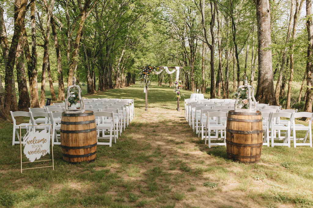 Wooden barrels and a welcome sign frame the dirt aisle toward Shay & Jacob’s floral arbor under Covey Creek’s trees.