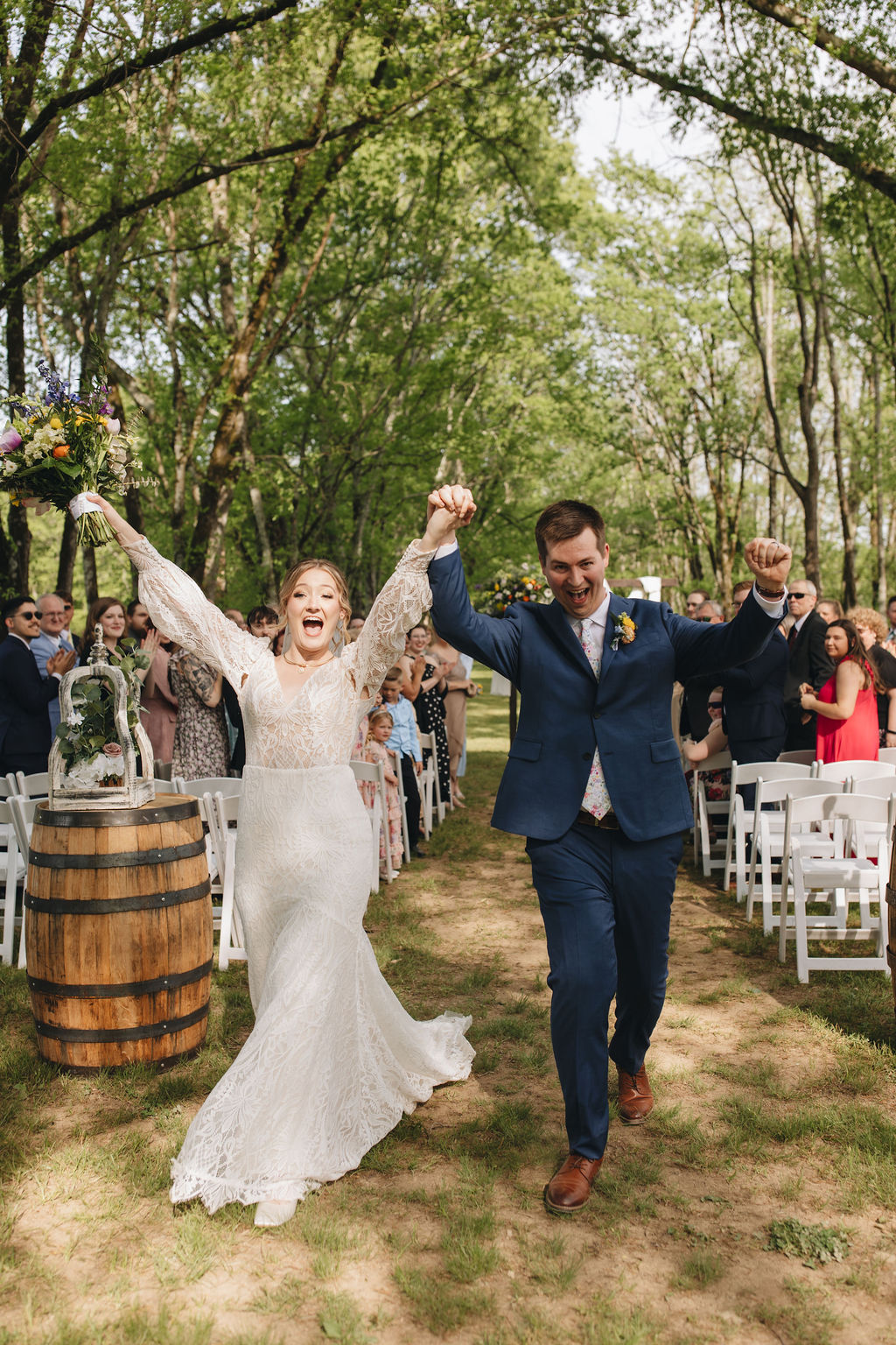 Shay & Jacob cheer down the tree-lined aisle at Covey Creek, bouquet raised as guests clap from white chairs.
