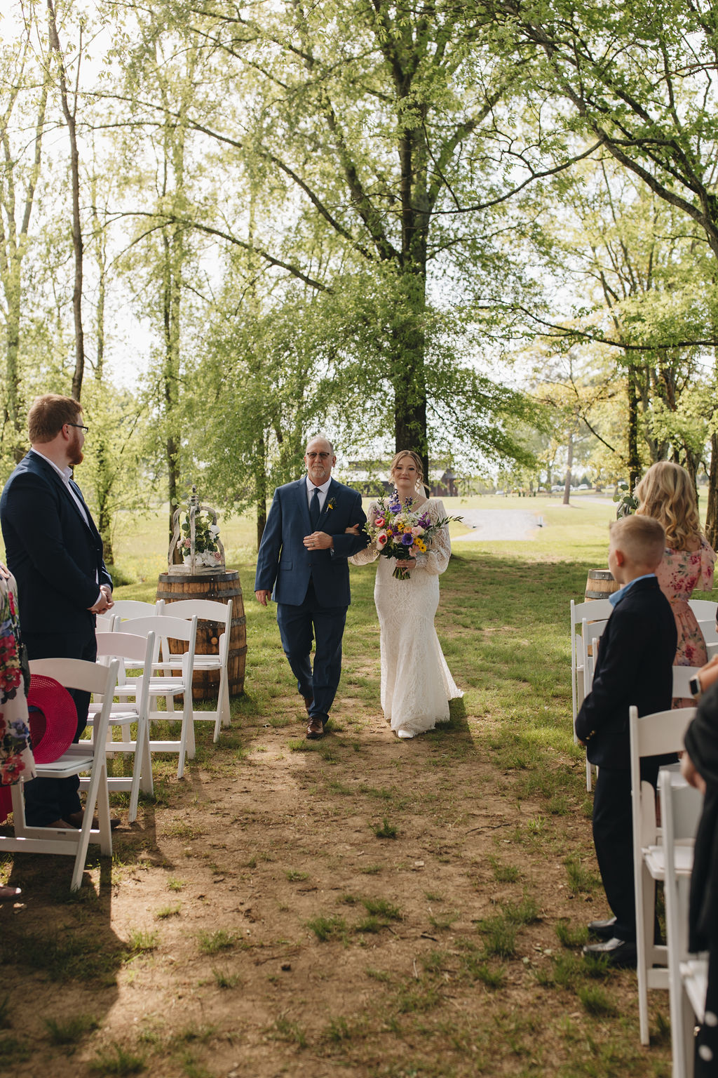 Shay walks the dirt aisle with her father, colorful bouquet in hand, as Jacob waits under the trees at Covey Creek.