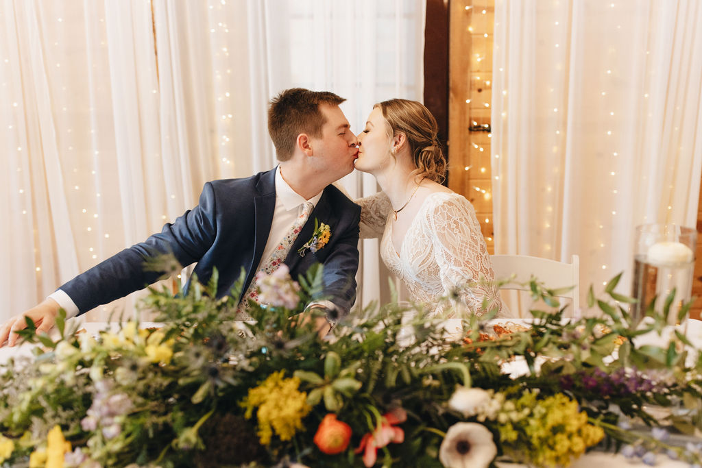Shay & Jacob kiss across their sweetheart table under fairy lights at Covey Creek, wildflower garland between them.