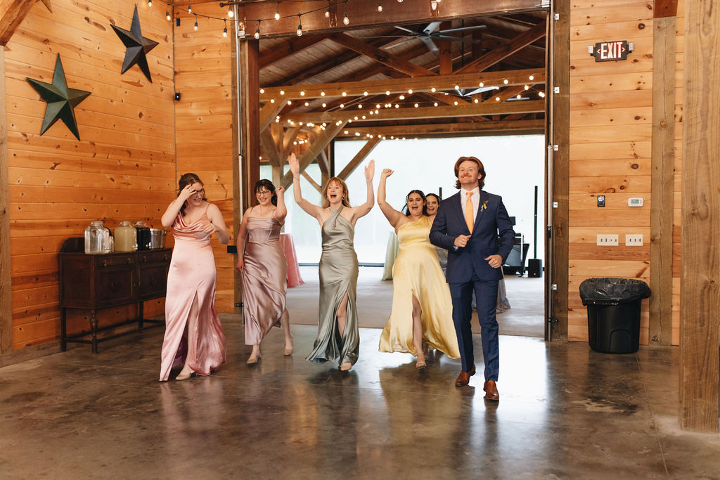 Bridesmaids in pastel satin cheer into Covey Creek’s barn at Shay & Jacob’s wedding, navy suits and string lights ahead.
