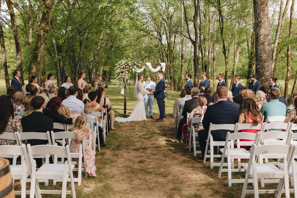 Guests face Shay & Jacob exchanging vows under a floral wooden arbor in Covey Creek’s tree-lined outdoor ceremony.