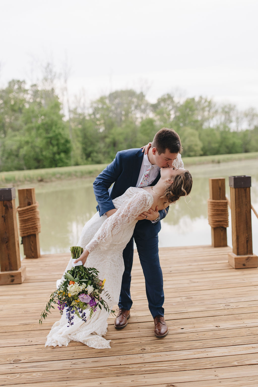 Jacob dips Shay for a kiss on the wooden dock by Covey Creek’s pond, her colorful wildflower bouquet in hand.