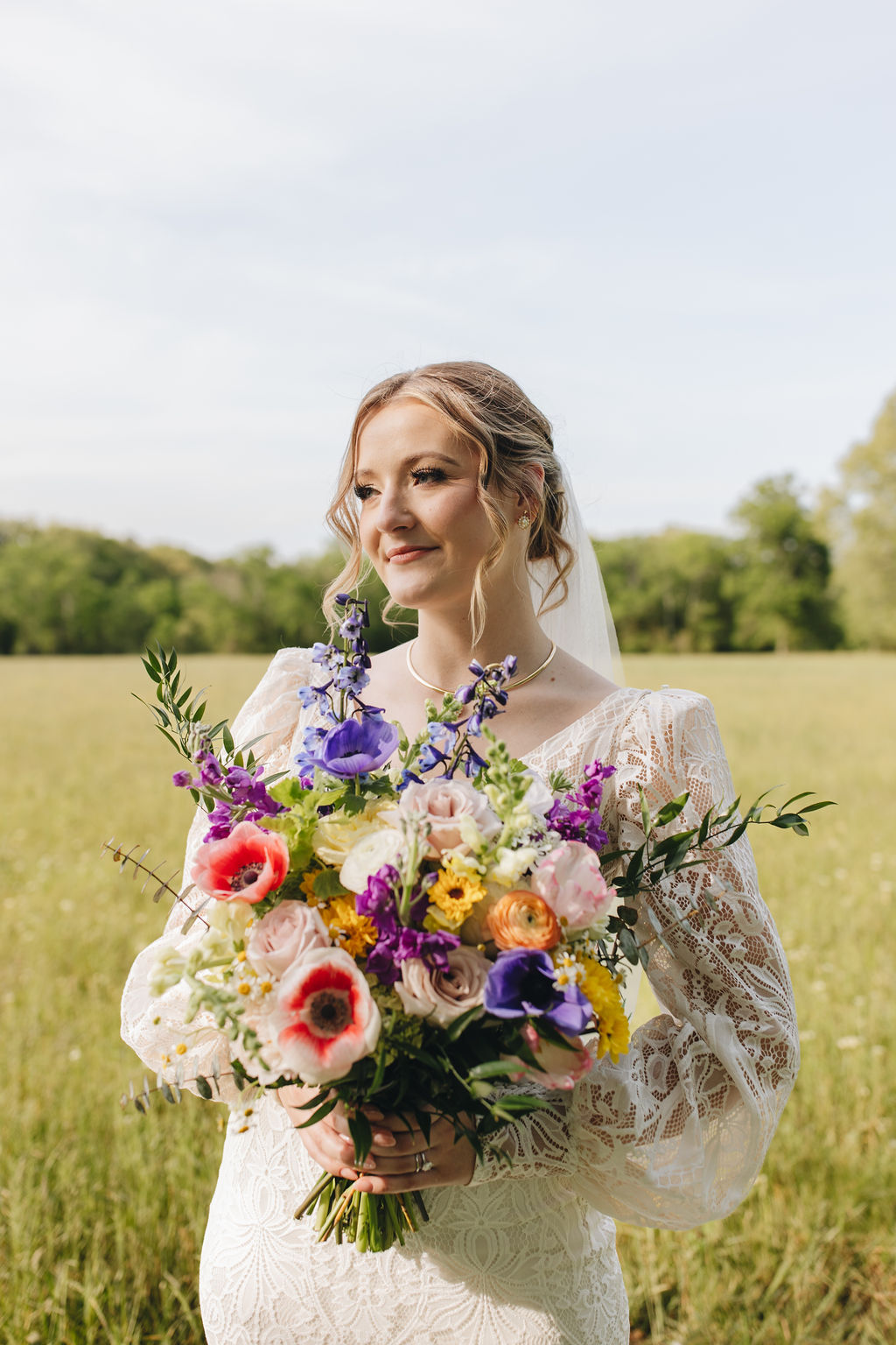 Shay holds a vibrant purple-and-yellow bouquet in a sunlit field at Covey Creek, lace sleeves and soft veil catching light.
