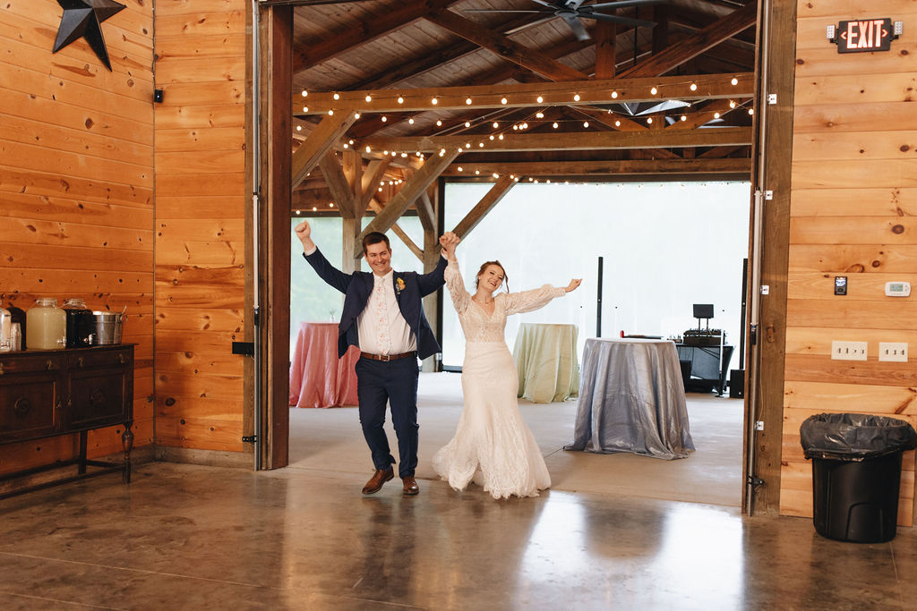 Shay & Jacob raise their arms entering Covey Creek’s barn reception, string lights and pastel cocktail tables behind them.