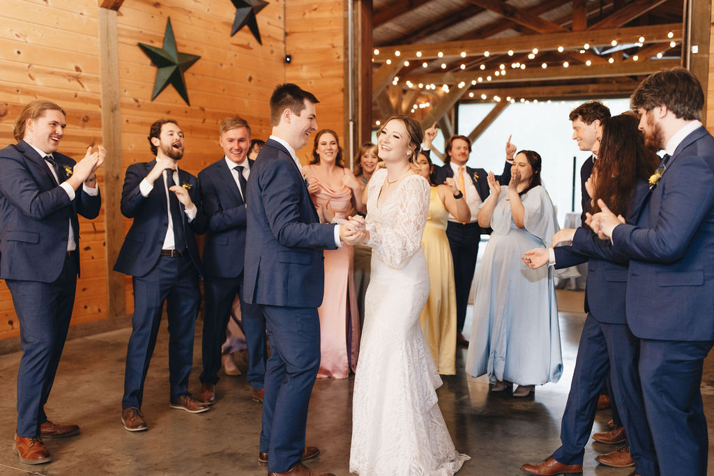 Shay & Jacob smile through cheering guests in Covey Creek’s barn reception, string lights on wood beams overhead.