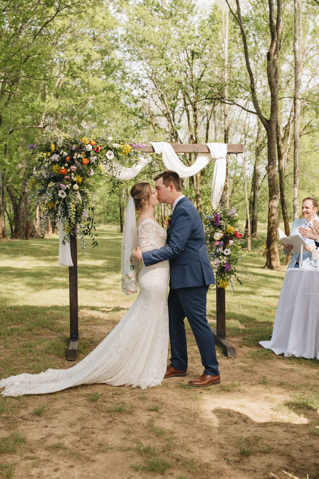 Shay & Jacob share their first kiss under a floral wooden arbor at Covey Creek, her embroidered veil trailing on grass.