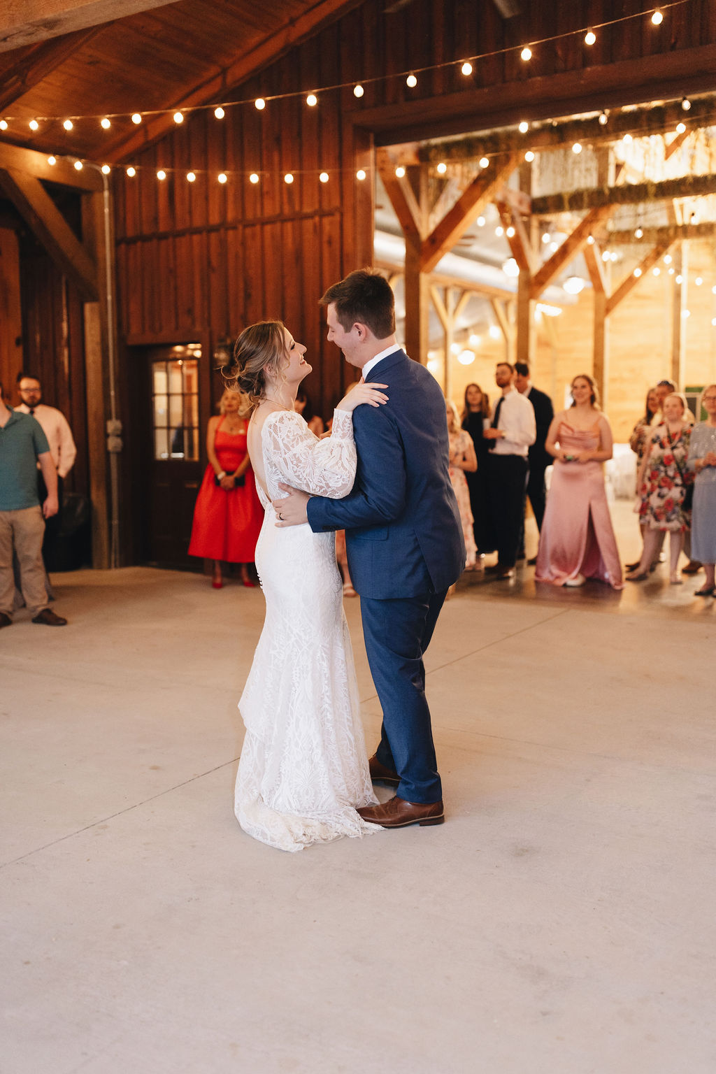 Shay & Jacob share their first dance under string lights in Covey Creek’s barn reception, guests watching from behind.