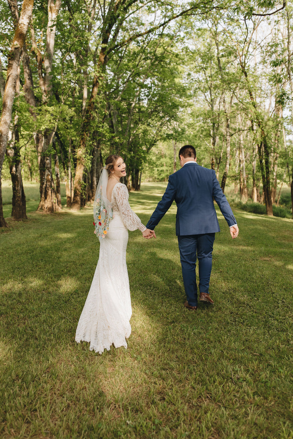 Shay looks back smiling as she and Jacob walk toward the tree grove at Covey Creek, floral veil trailing.