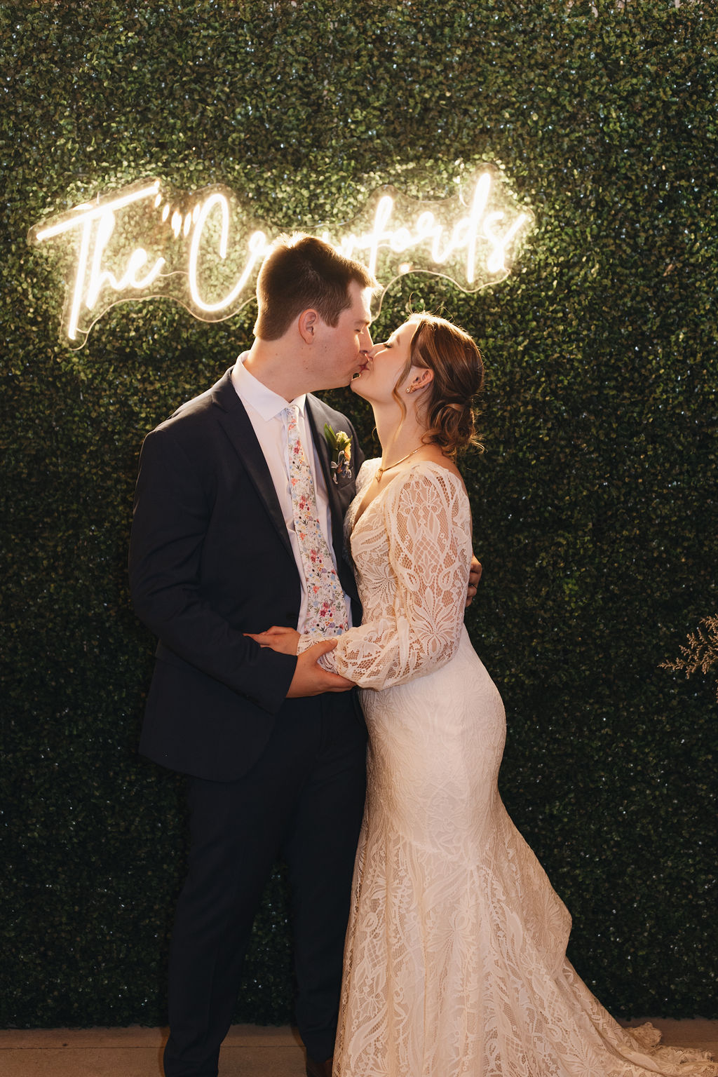 Shay & Jacob kiss under a glowing “The Crawfords” neon sign against a green hedge wall at Covey Creek.
