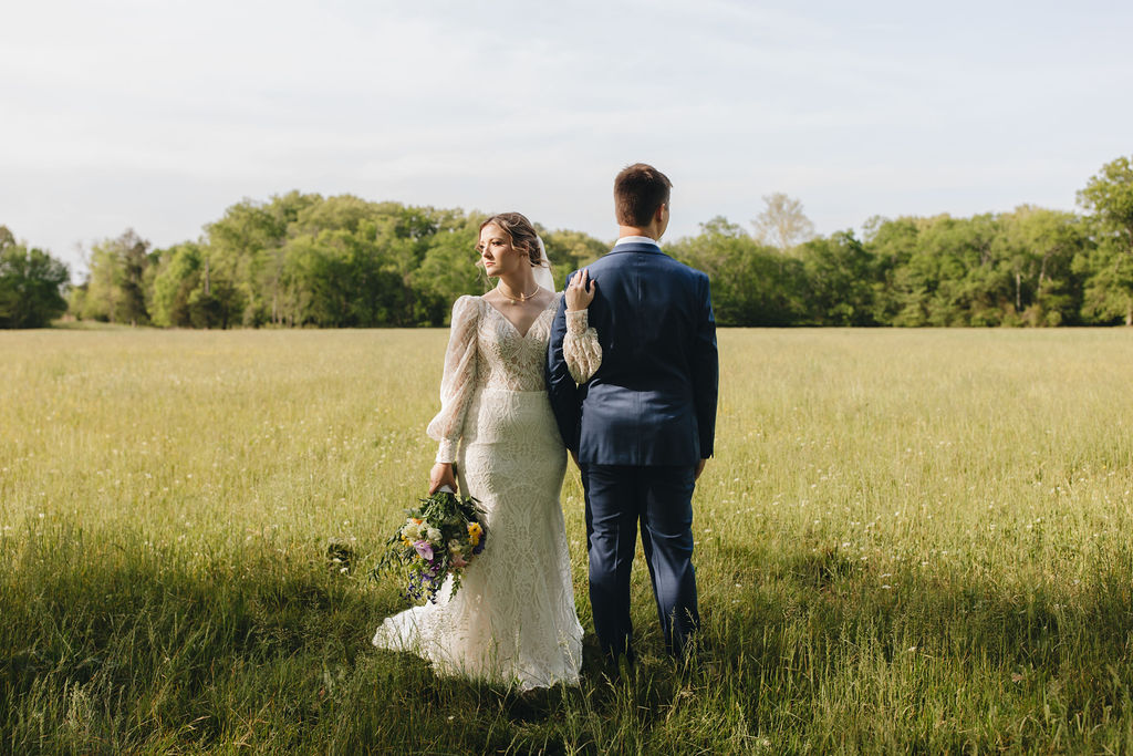 Shay rests a hand on Jacob’s shoulder in a tall-grass field at Covey Creek, colorful bouquet at her side.