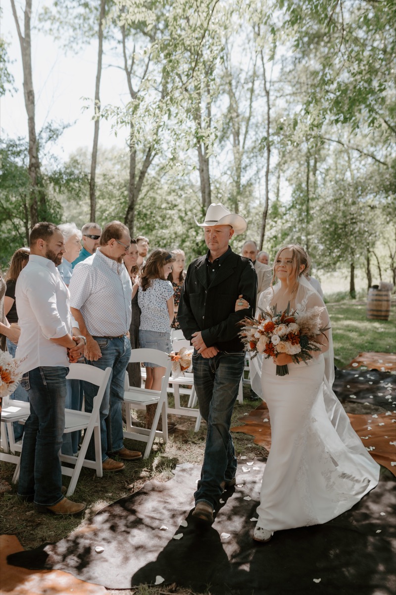 Shay walks a petal-strewn cowhide aisle under trees at Covey Creek for Shay & Wyatt's outdoor ceremony.