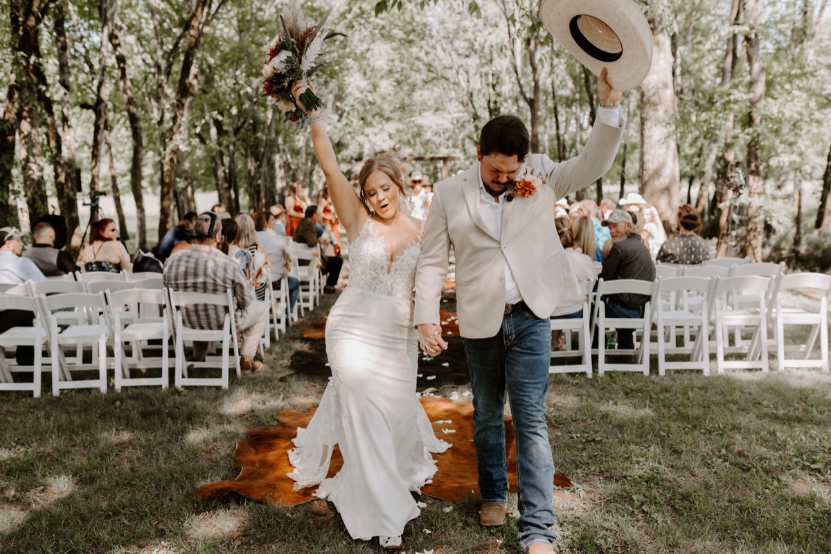 Shay & Wyatt recess down the cowhide aisle at Covey Creek, bouquet and cowboy hat raised in celebration.
