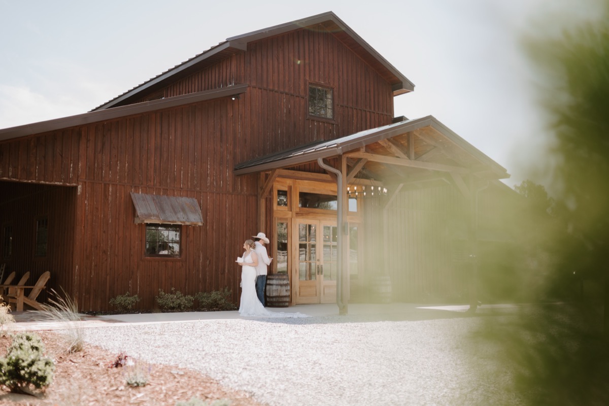 Shay & Wyatt stand close outside the Covey Creek barn, her gown trailing gravel as he wears a white cowboy hat.