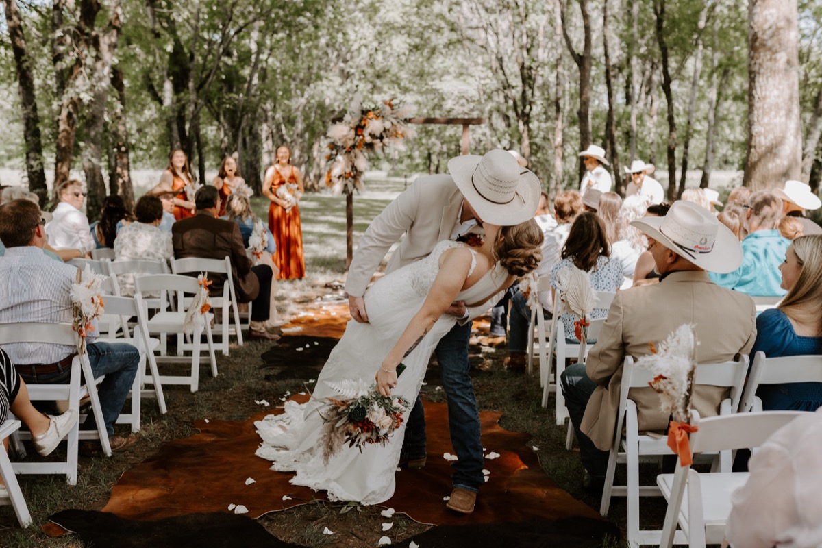 Shay & Wyatt share a dip kiss on the cowhide aisle under a floral arbor as guests watch at Covey Creek.